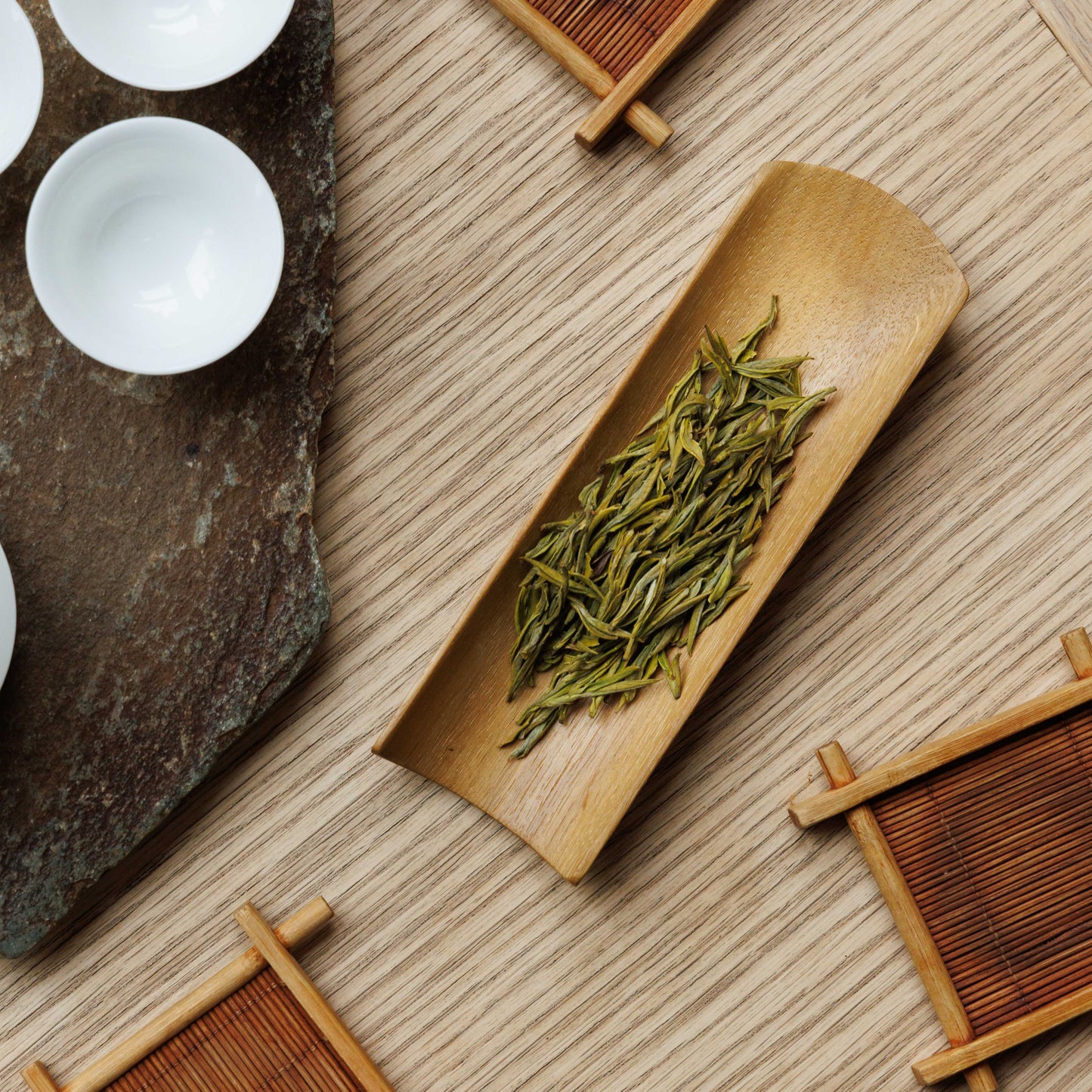 A wooden tray containing rolled green tea leaves, with white cups and tea-making equipment in the background.